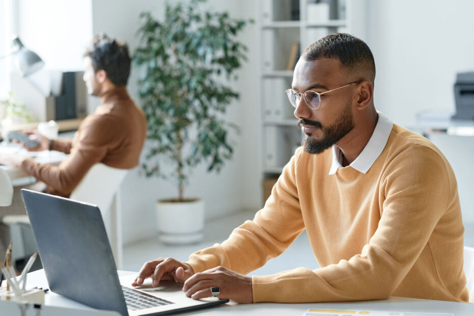 Serious young mixed race businessman in yellow sweater working with website on laptop in modern office | ANG