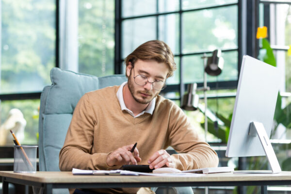 Accounting, audit. A young man works with documents and bills in the office. Calculates and checks reporting, balance, and budget on a calculator.
