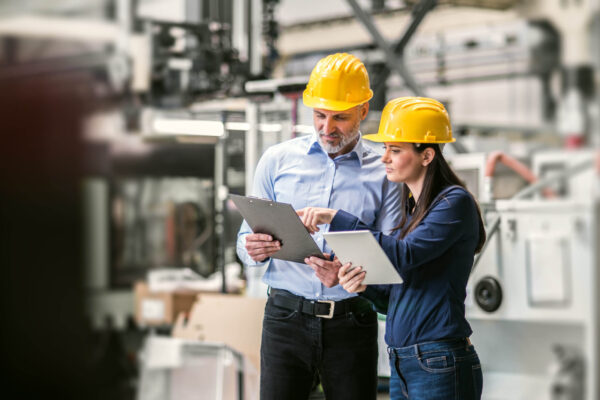 Two project managers standing in modern industrial factory. Manufacturing facility with robotics, robotic arms and automation.