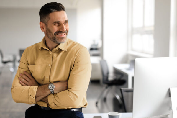 Happy middle aged businessman standing by his workplace, looking aside at copy space, thinking of new startup or project, smiling at good thoughts, planning investment, enjoying success