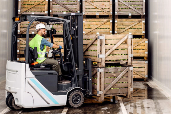 Modern-warehouse,-work-on-forklift,-industry-and-organic-agribusiness.-Smiling-millennial-man-in-helmet-in-forklift-lifts-up-green-apples-in-container-on-shelves-with-many-wooden-crates