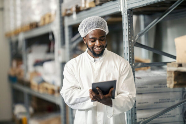 Storage-manager-in-sterile-uniform-working-on-checklist-and-smiling-at-tablet-at-warehouse